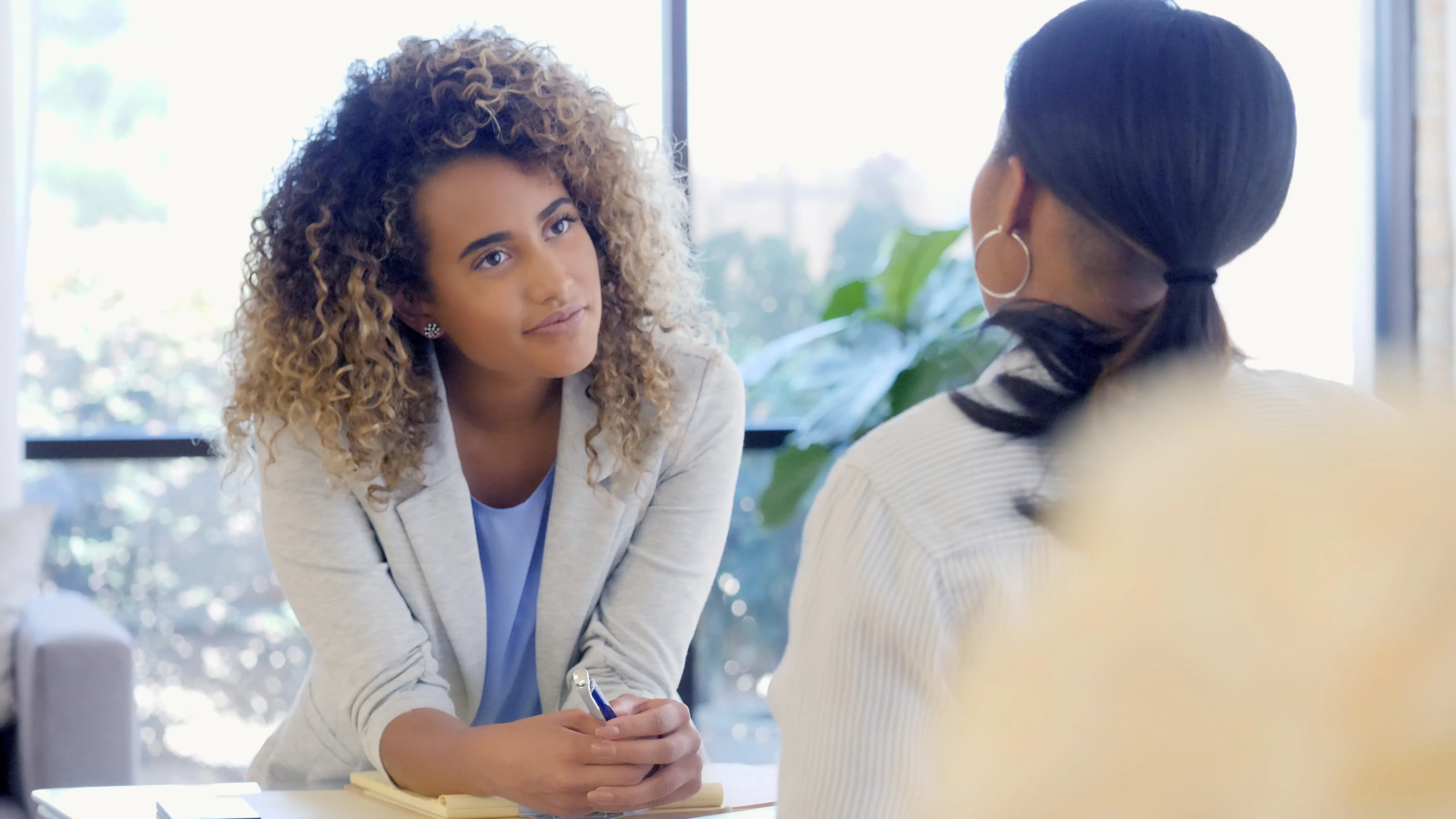 A diverse group of professionals in business attire stack their hands together in a team huddle over a desk, symbolizing unity and collaboration. A printed document and eyeglasses rest on the desk beneath their hands.