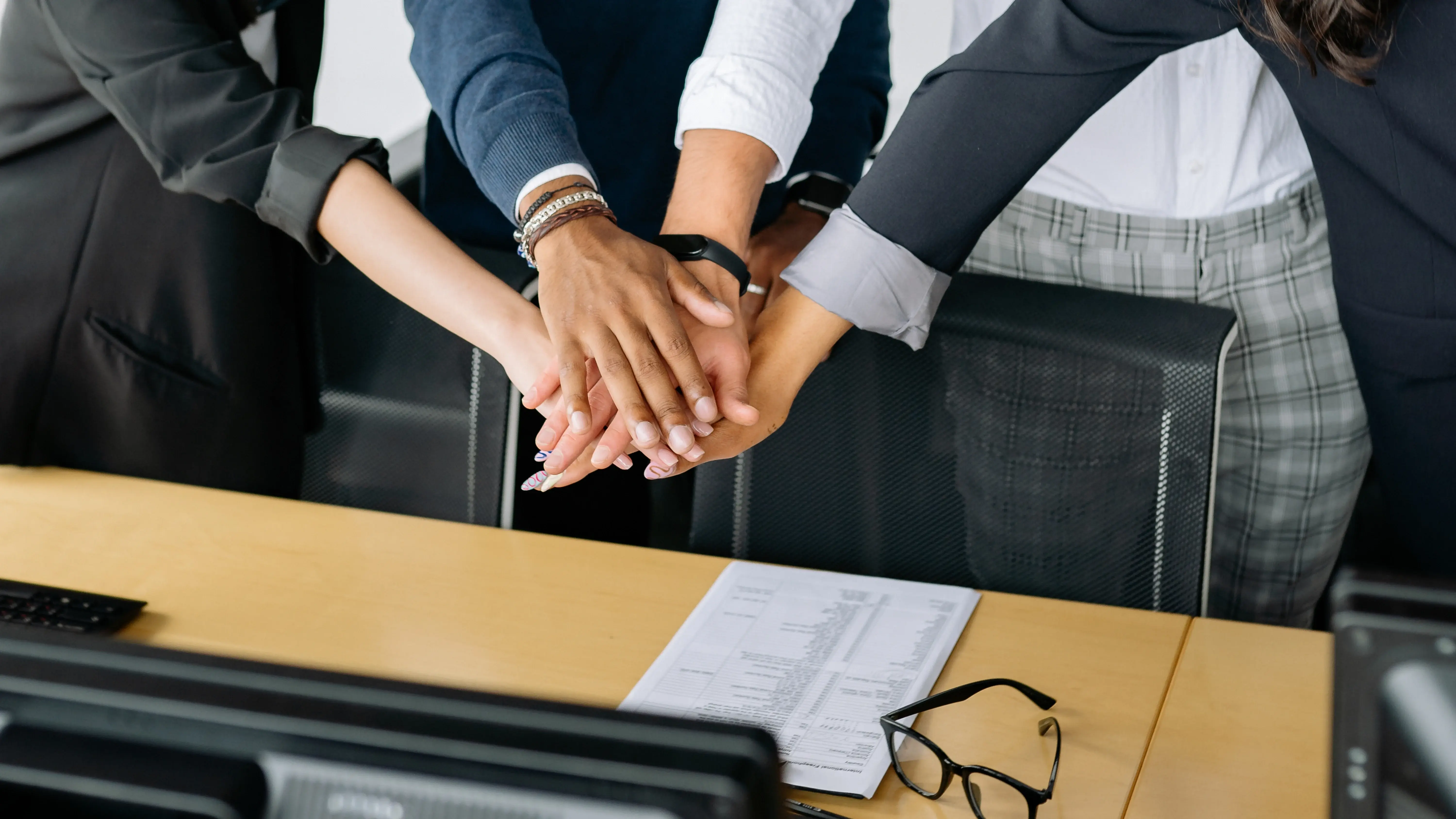 A diverse group of professionals in business attire stack their hands together in a team huddle over a desk, symbolizing unity and collaboration. A printed document and eyeglasses rest on the desk beneath their hands.